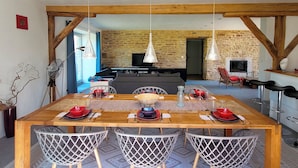 A dining area with a wooden table set for four, surrounded by metal chairs. A stone wall and wooden beams are visible in the background.