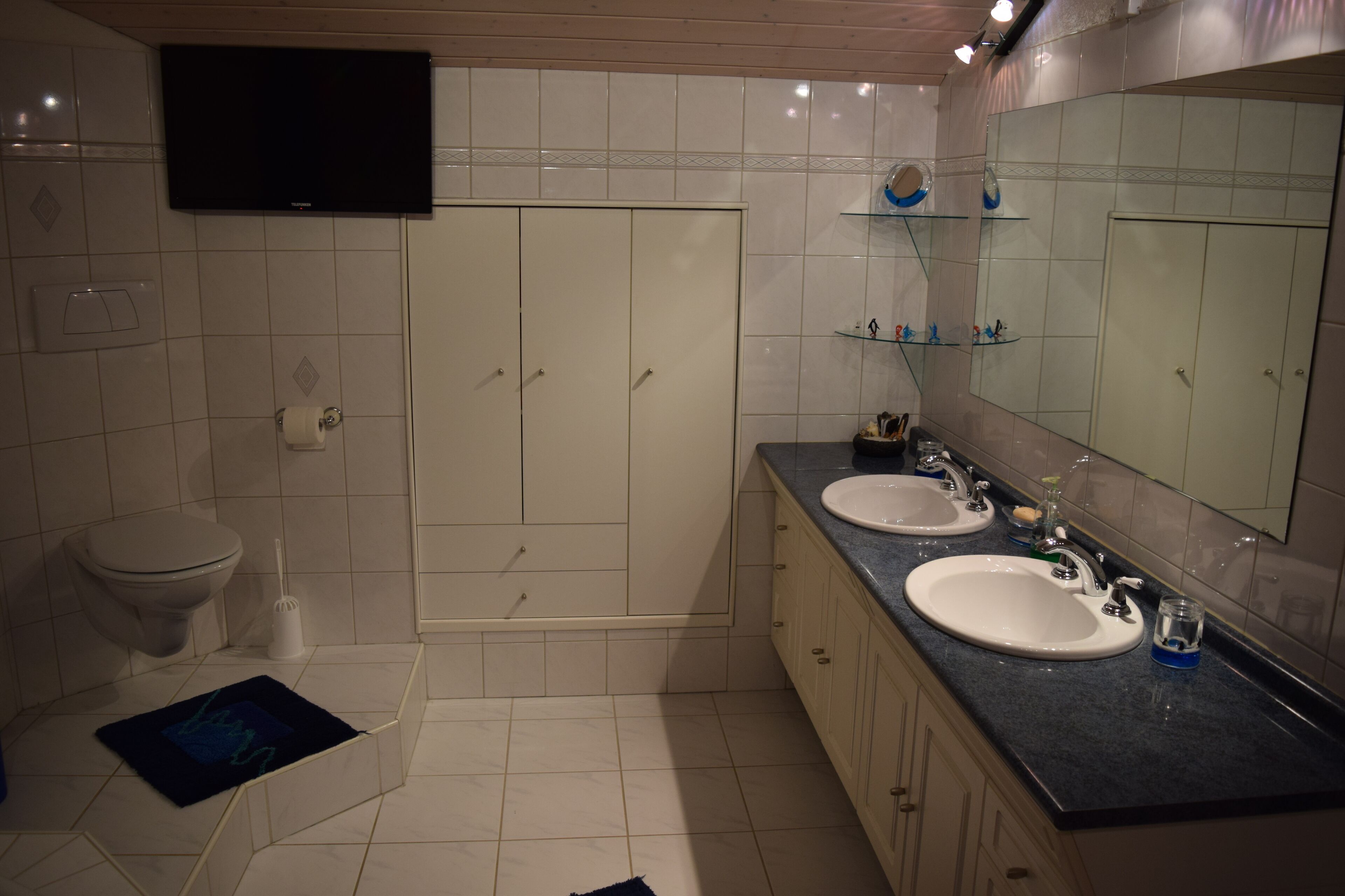 Et moderne badeværelse med to håndvaske, toilet, brusebad og et ophængt fjernsyn.