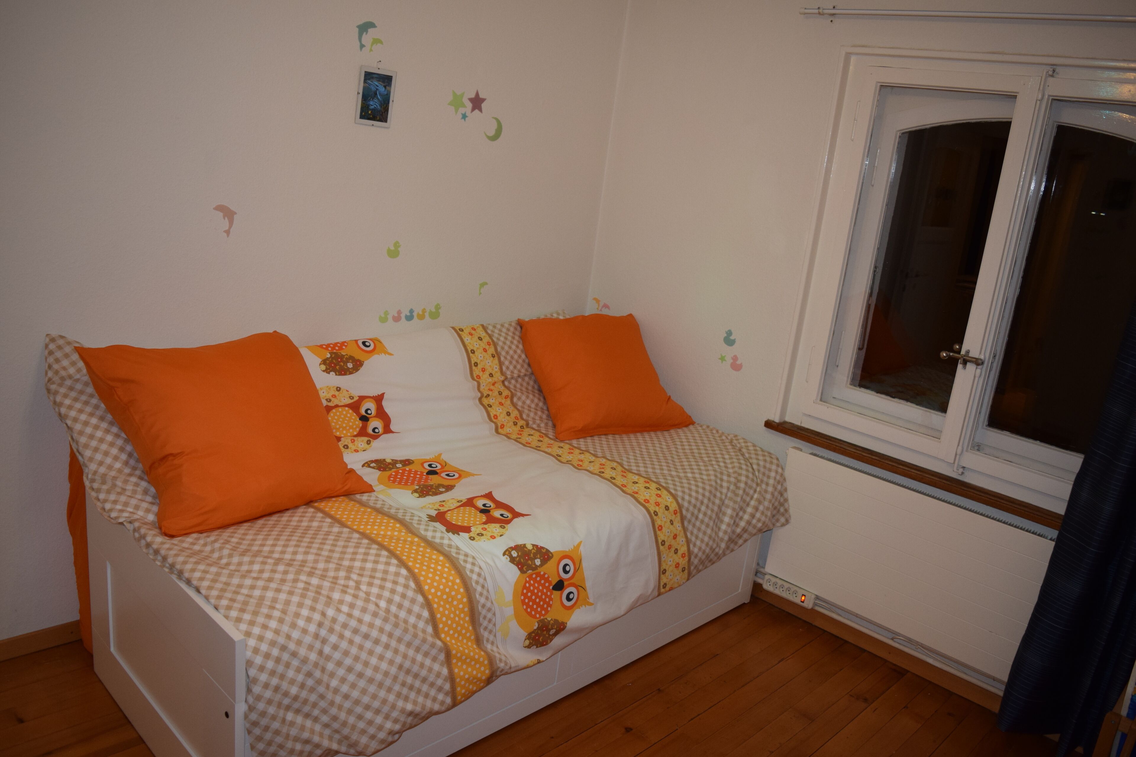 Une petite chambre à coucher accueillante, avec un lit simple agrémenté de draps orange et blancs, une fenêtre offrant une belle vue et un mur décoré d’autocollants représentant des étoiles et une lune.