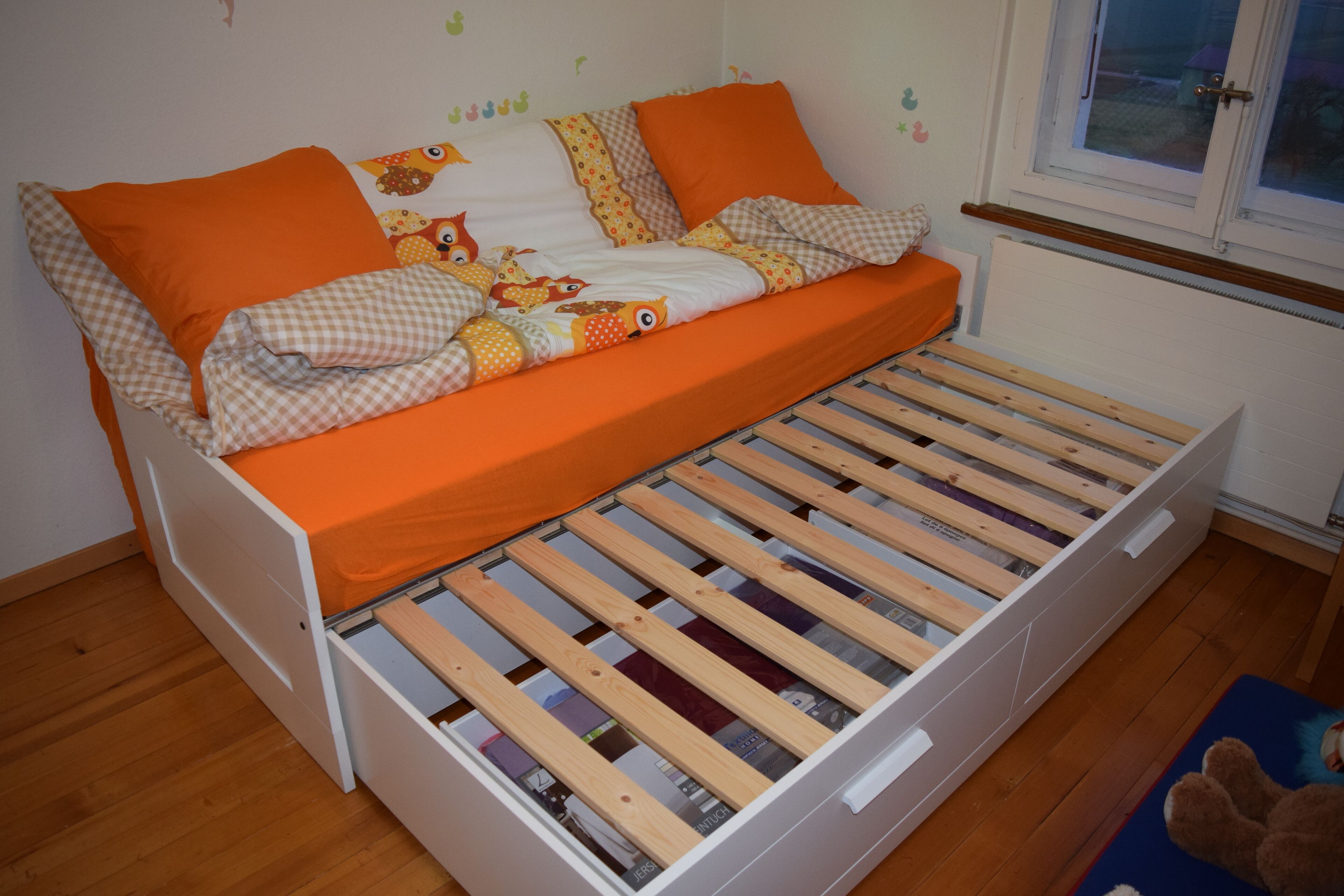 Een eenpersoonskamer met een houten bedframe, oranje beddengoed en een raam met uitzicht op buiten.