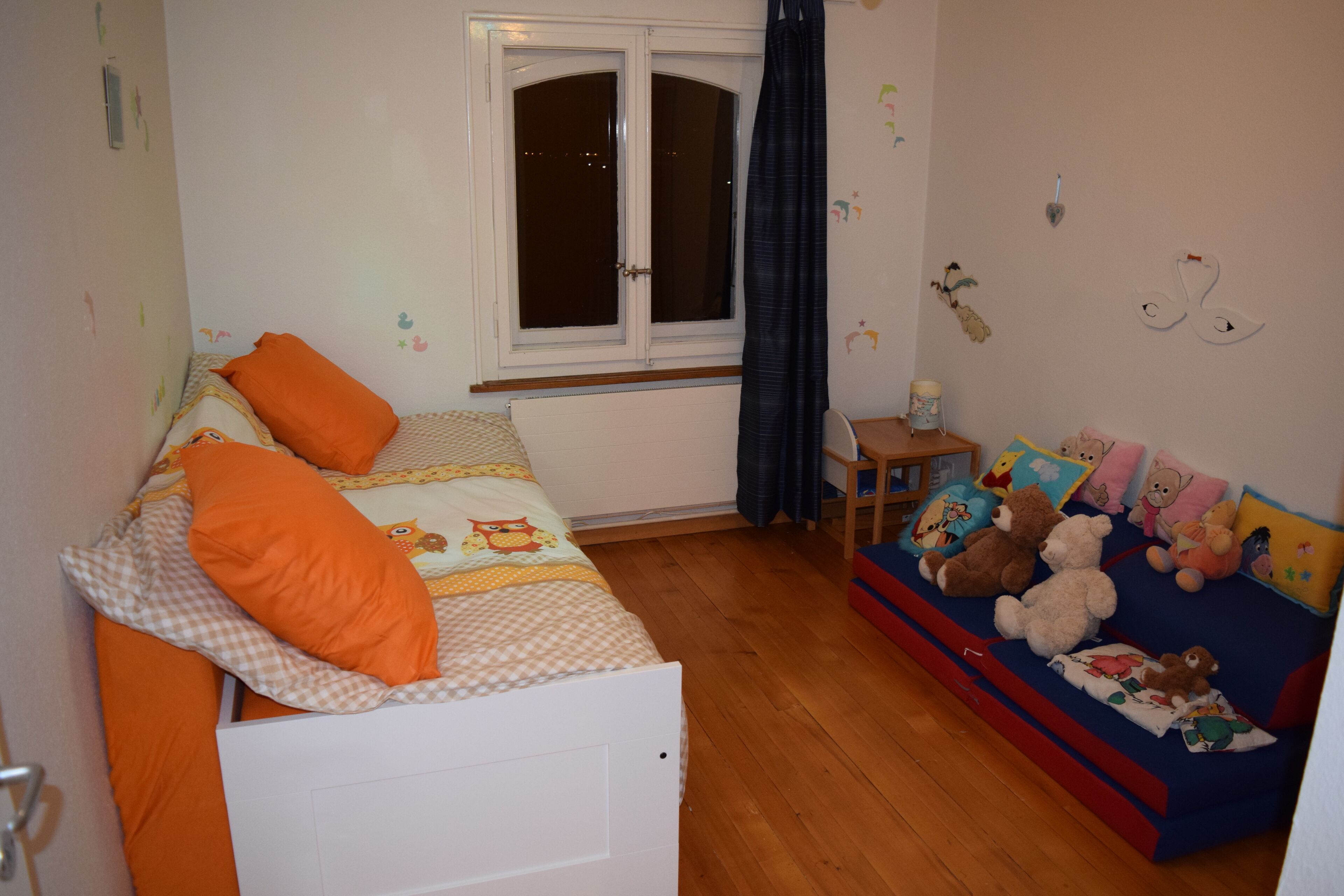 Ein Schlafzimmer mit einem Bett, einem Fenster, einem kleinen Tisch und einer Couch mit Kuscheltieren.
