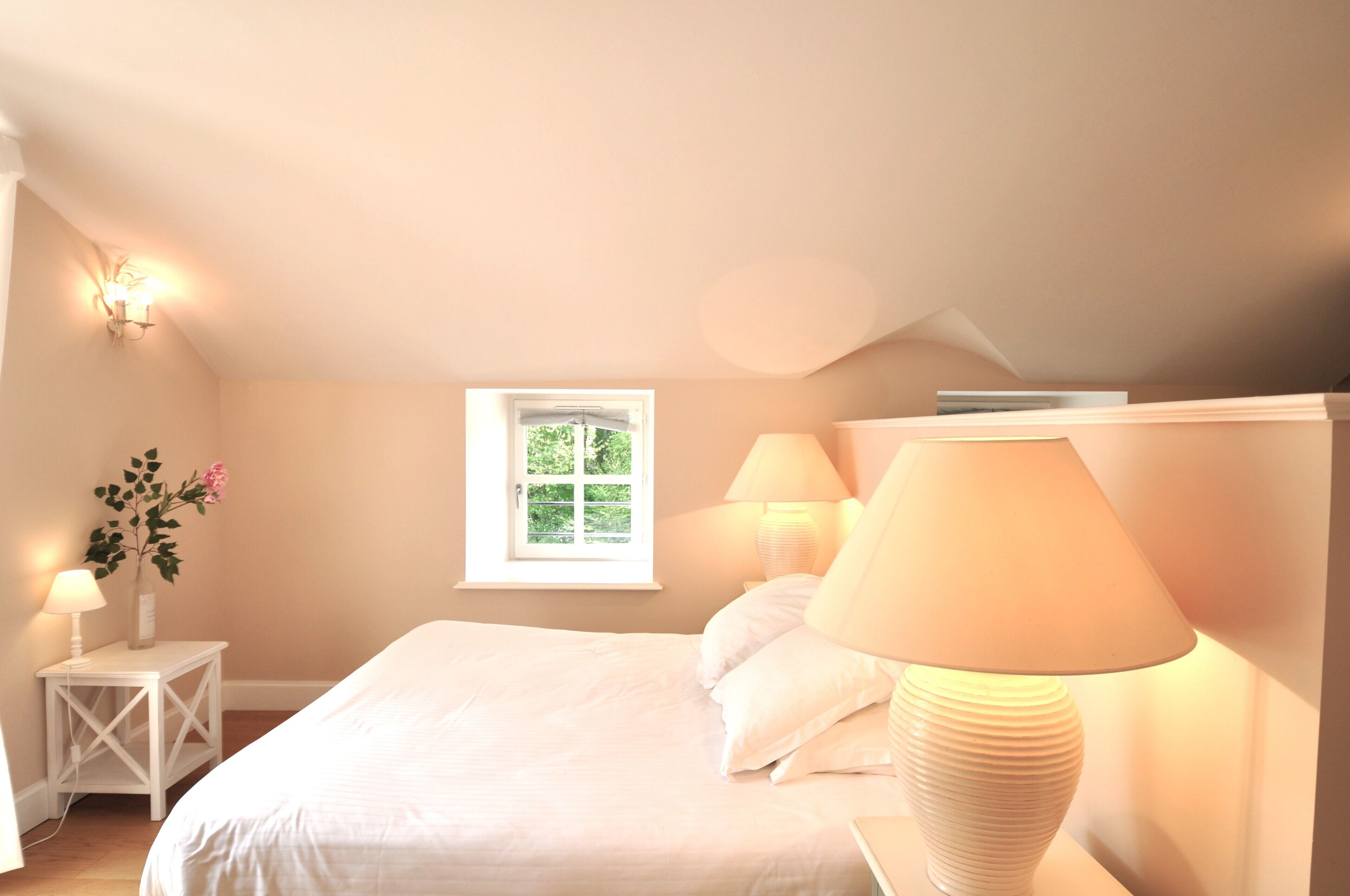 Une chambre à coucher avec un lit, deux lampes de chevet, une table de chevet avec une lampe, un vase de fleurs et une fenêtre donnant sur un paysage verdoyant.