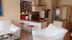 Ein modernes Wohnzimmer mit einem weißen Sofa, zwei roten Barhocker, einem Glastisch und einer Küchenzeile mit Herd, Backofen und Mikrowelle.