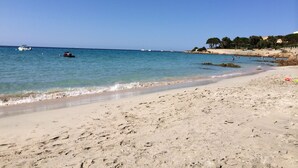 Ein Strand mit klarem, blauem Wasser, sandigem Ufer und Booten in der Ferne.