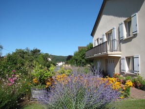 Un bâtiment à deux étages doté d’un balcon, entouré d’un jardin où poussent différentes sortes de fleurs et de plantes.