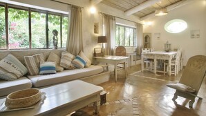 Ein gemütliches Zimmer mit einer Couch, Esstisch und Stühlen, großen Fenstern mit Vorhängen und einer Holzbalkendecke.