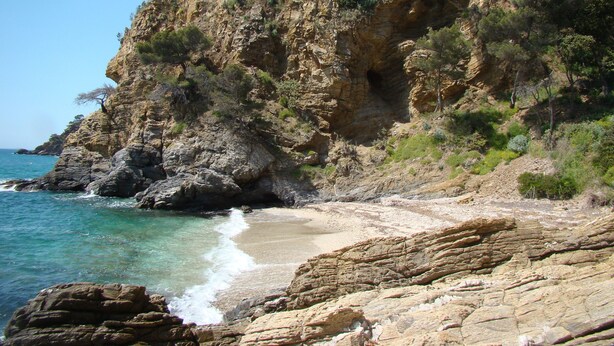 Eine felsige Küste mit klarem, blauem Wasser, einem Sandstrand und einer steilen Klippe mit spärlicher Vegetation.