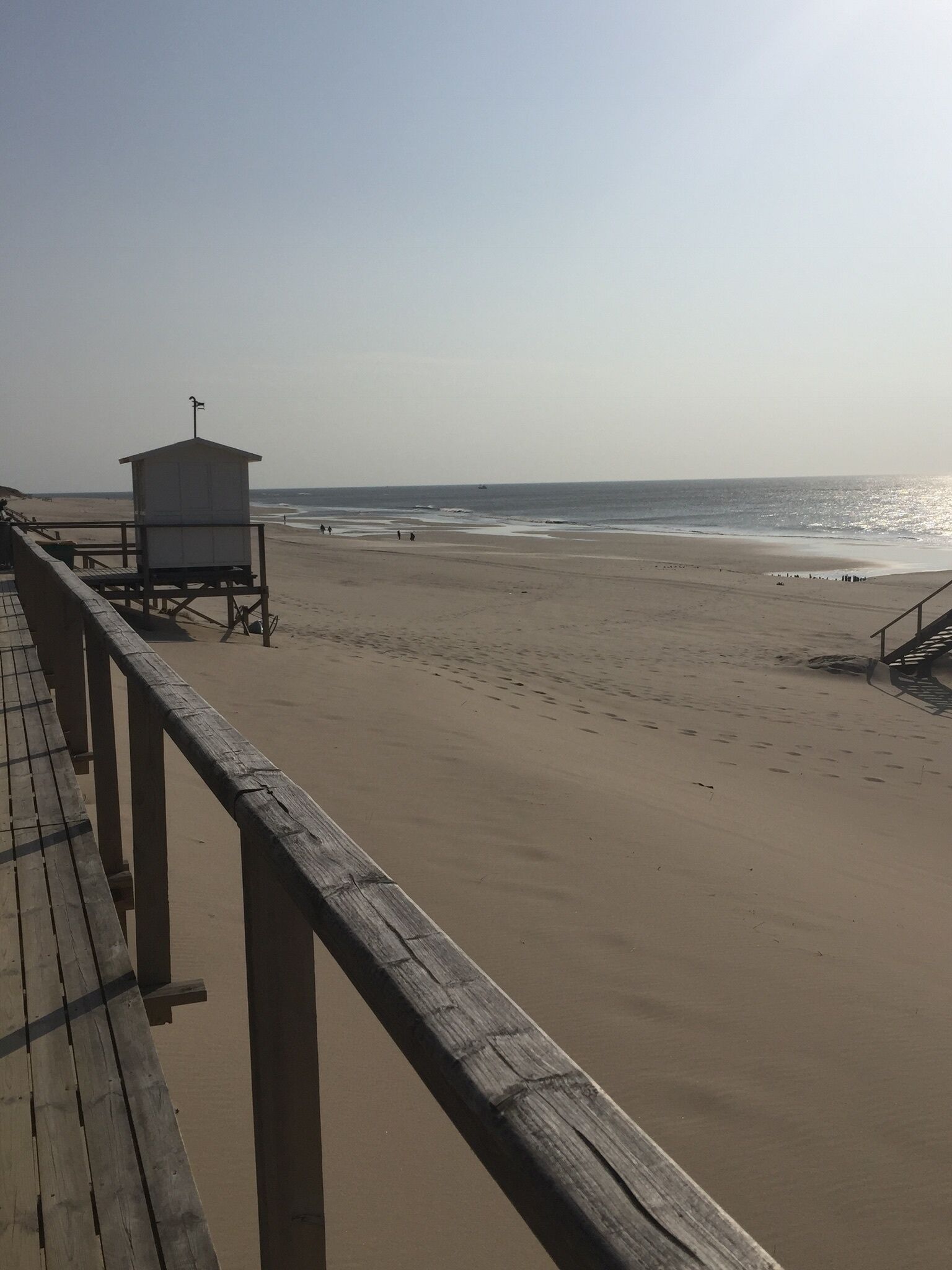 Une promenade en bois mène à un poste de surveillance des sauveteurs, qui surplombe une plage de sable avec l’océan en arrière-plan.