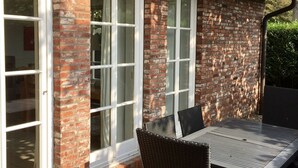 A brick house with a tiled roof, white-framed windows, and a patio with a table and chairs.