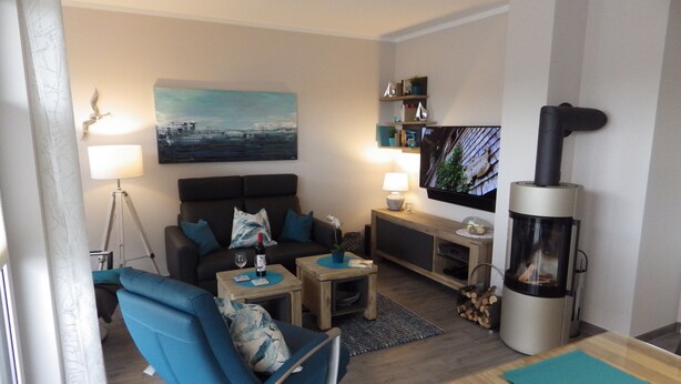 Een moderne woonkamer met een bank, een blauwe fauteuil, een salontafel, een televisie, een open haard en een schilderij aan de muur.