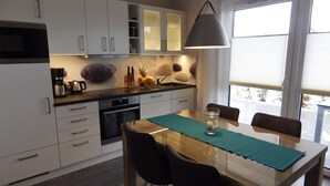 Een moderne keuken met witte kastjes, een zwarte magnetron en een koffiezetapparaat. Er staat een eettafel met stoelen en een glas water op tafel.