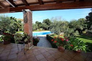 A covered patio with a view of a swimming pool, surrounded by potted plants and a brick pillar.