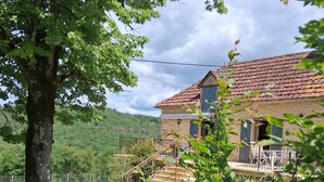 Une maison de campagne au toit recouvert de tuiles, un escalier menant à un porche et un grand arbre au premier plan.