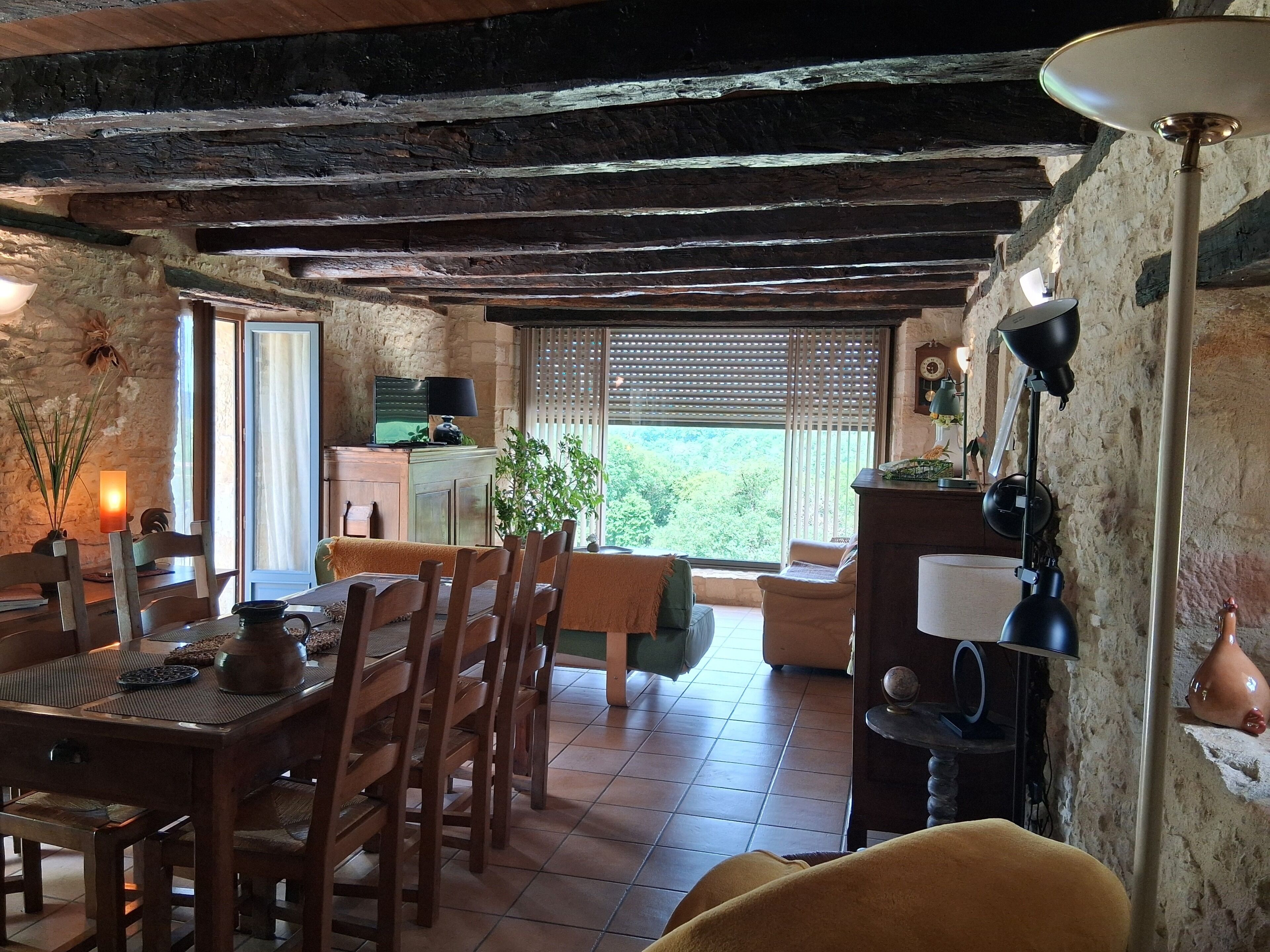 Un intérieur rustique avec des poutres apparentes, un coin repas avec des meubles en bois, une cuisine dotée d’une cuisinière moderne et une grande fenêtre avec des stores.
