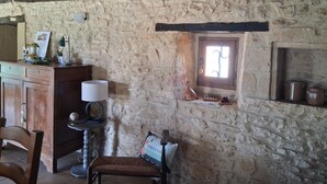 Une pièce rustique aux murs de pierre, avec un bureau en bois, une chaise, une petite fenêtre et une étagère où sont posés des pots.
