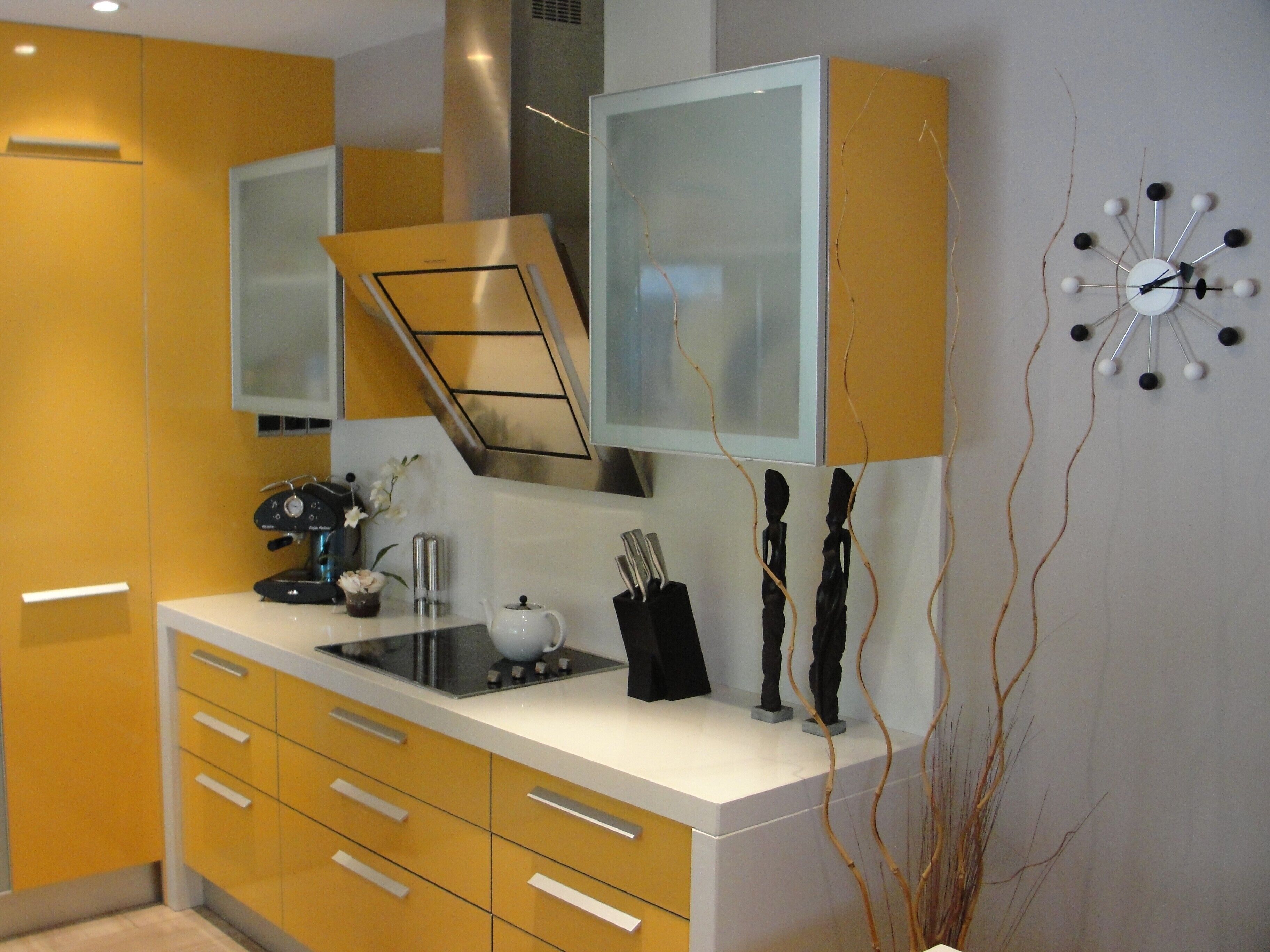 A modern kitchen with yellow cabinets, a white countertop, and a unique wall clock.