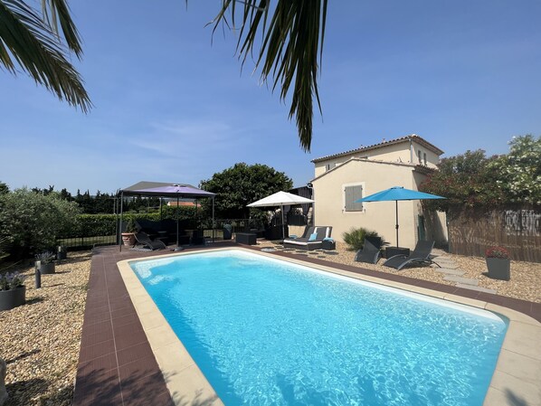 Une piscine rectangulaire dont le bord est revêtu de carrelage, entourée d’un espace patio aménagé avec des chaises longues et des parasols.