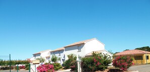 A residential entrance with a paved driveway, a white building with a red-tiled roof, and pink flowering bushes.