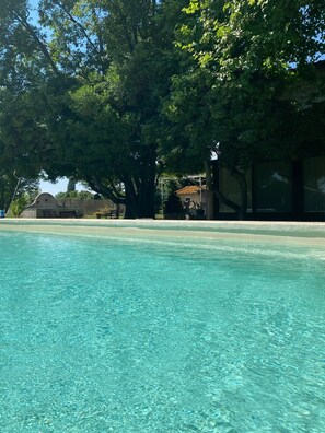 A clear swimming pool with a tiled edge, surrounded by lush green trees and a building with a tiled roof in the background. - Provencal farmhouse in the countryside Arlesienne - Heated pool (Arles)