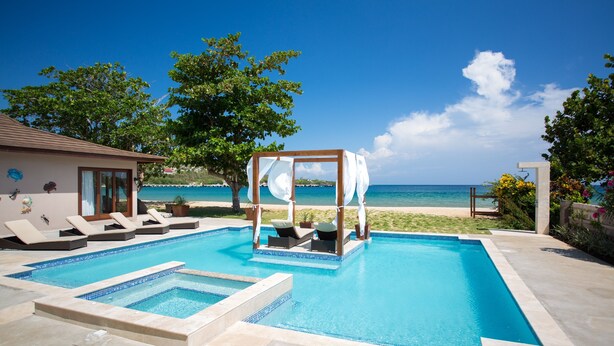 Una piscina rectangular con un pabellón de madera en el centro, rodeada de sillas de descanso y vista a la playa al fondo.
