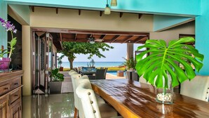 Un comedor con mesa y sillas de madera, vista a la playa y una planta tropical.
