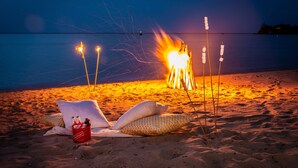 Escena de playa con una hoguera, un cubo rojo con botellas y una silla de salón con almohadones.