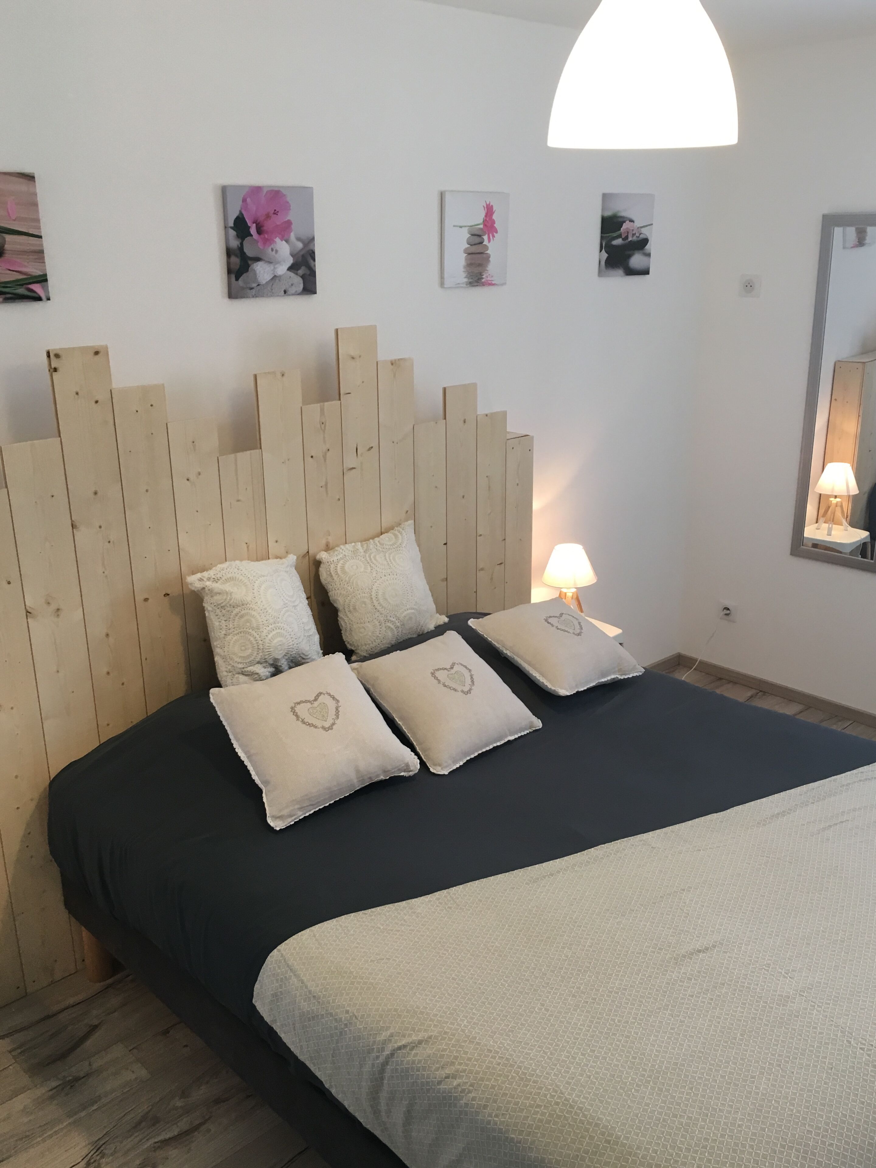 Ein Schlafzimmer mit einem hölzernen Kopfteil, einer schwarzen Tagesdecke und vier Kissen mit Herzmotiven. An den Wänden befinden sich Bilder und eine Nachttischlampe.