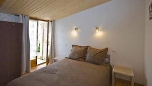 Un dormitorio con cama, mesita de noche y un taburete pequeño. Hay dos luces fijadas a la pared y una puerta corrediza de cristal que da a un balcón.