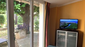 A room with a television on a cabinet, a sliding glass door leading to an outdoor area with trees and plants, and a small table. - House garden garage 10 mn downtown (Nîmes)