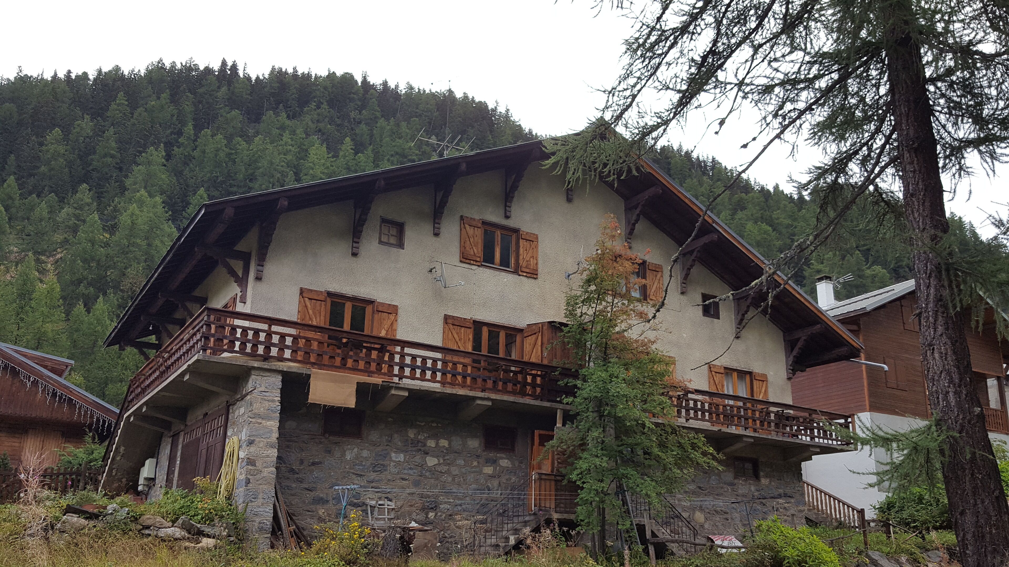 A two-story mountain lodge with stone walls, wooden balconies, and a forested backdrop.