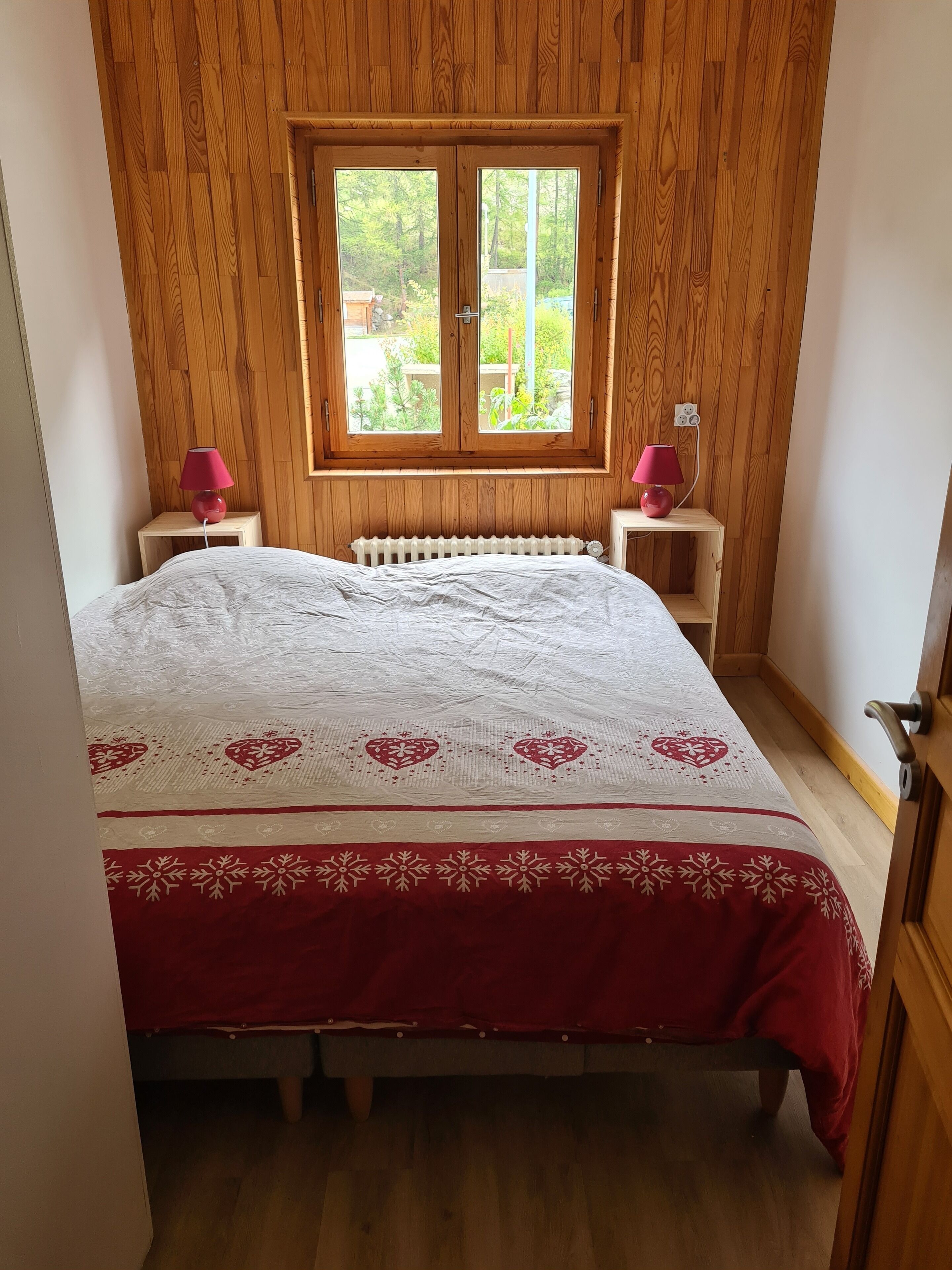 Un dormitorio con pared de madera, ventana con vista a vegetación, cama con colcha blanca y roja, y dos mesitas de noche con lámparas.