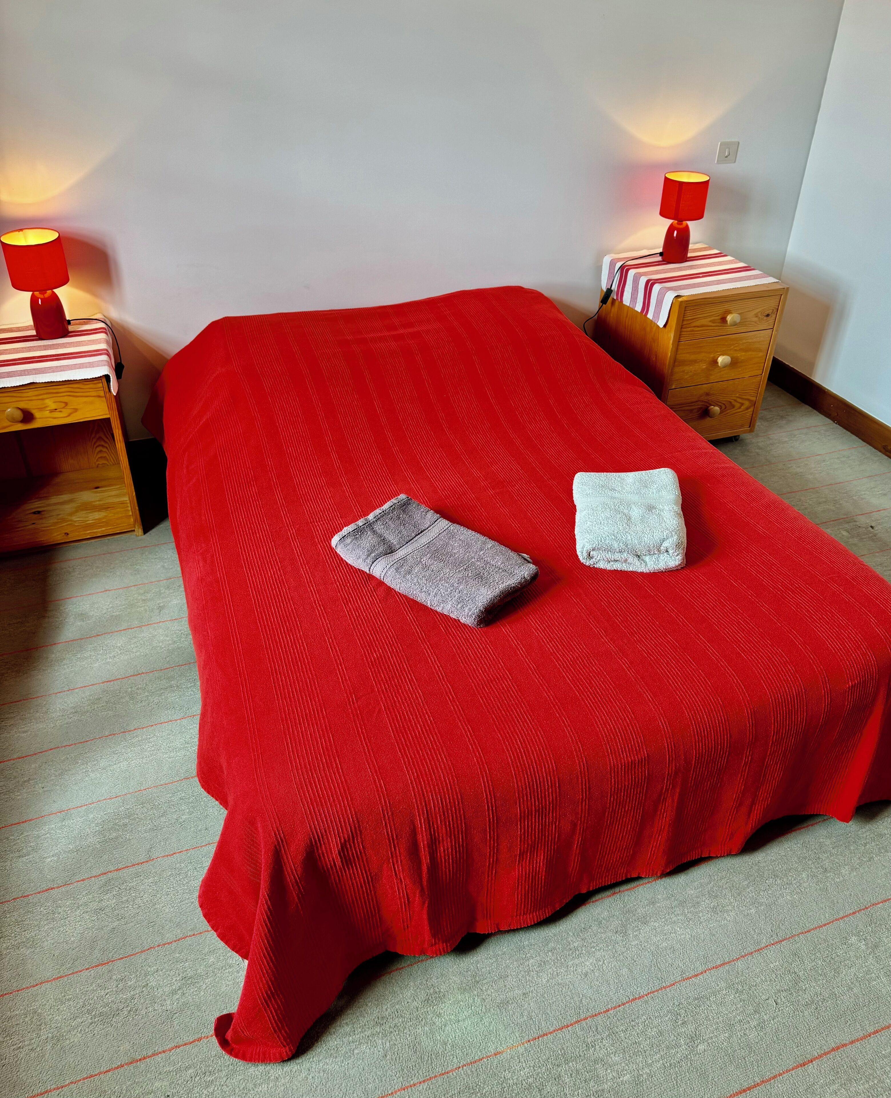 Un lit simple avec une couvre-lit rouge, deux oreillers, une table de chevet avec une lampe et une petite table en bois.