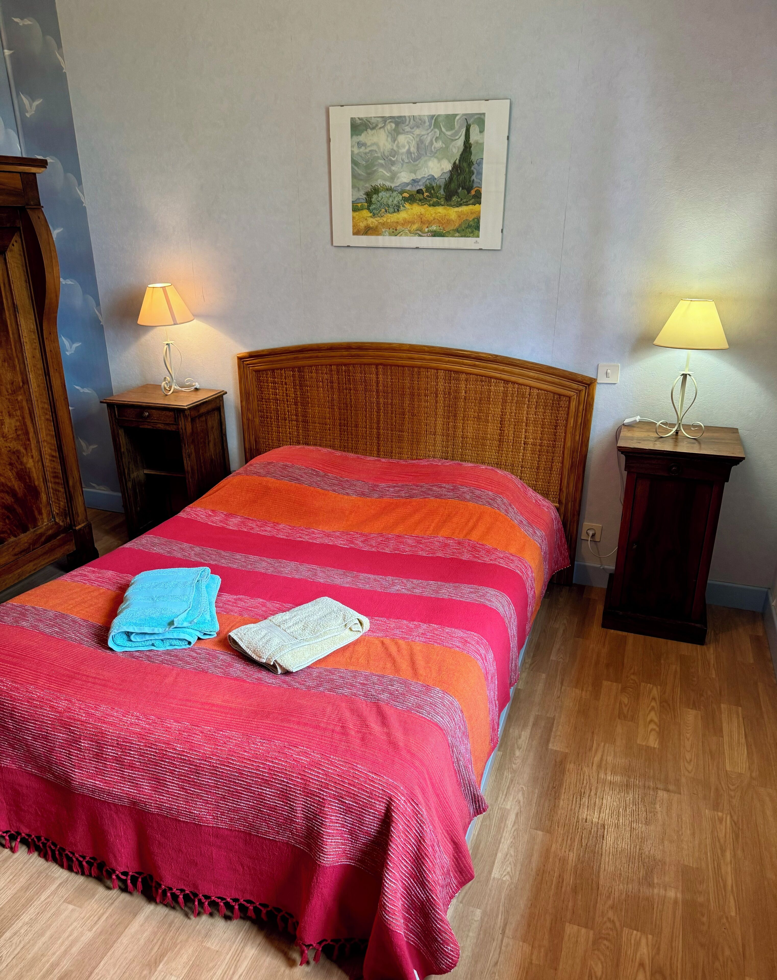 Una cama bien hecha con una colcha a rayas y dos toallas, dos lámparas de noche, un cabecero de madera, un cuadro enmarcado en la pared y suelo de madera.