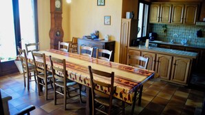 A dining area with a long table and chairs, a clock on the wall, and a kitchen area with cabinets and a sink.