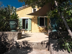 A rustic building with a tiled patio, a door with green shutters, and two large green planters.