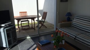 En stue med sofa, fjernsyn, bord og stol. En skydedør fører til et udendørs område med et bord og stole.