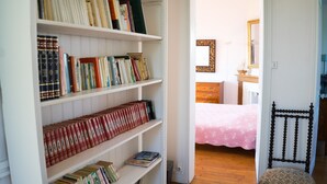 Una habitación con piso de madera, una cama con colcha rosa y una estantería llena de libros.
