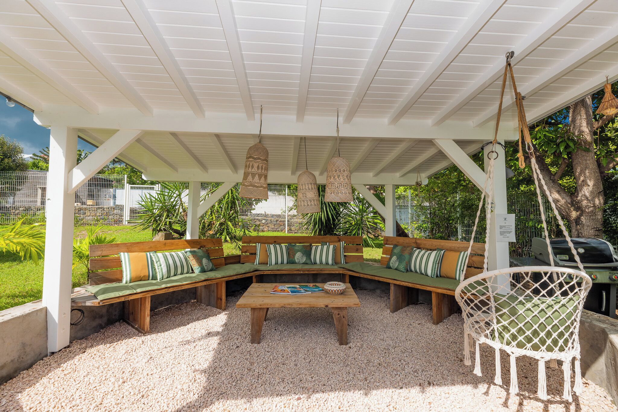 A covered outdoor seating area with a wooden bench and a hanging chair, surrounded by greenery.