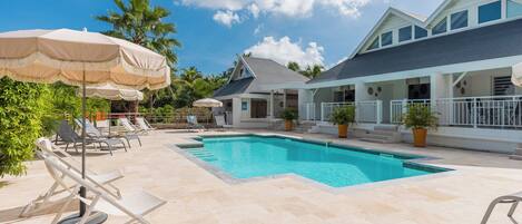 A swimming pool with lounge chairs and umbrellas, surrounded by a house with a balcony and potted plants.