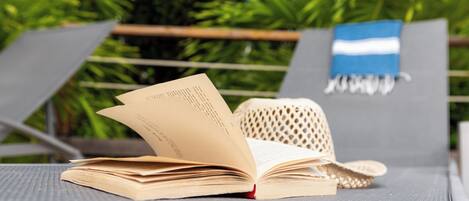 An open book on a lounge chair with a woven hat beside it, set against a backdrop of greenery.