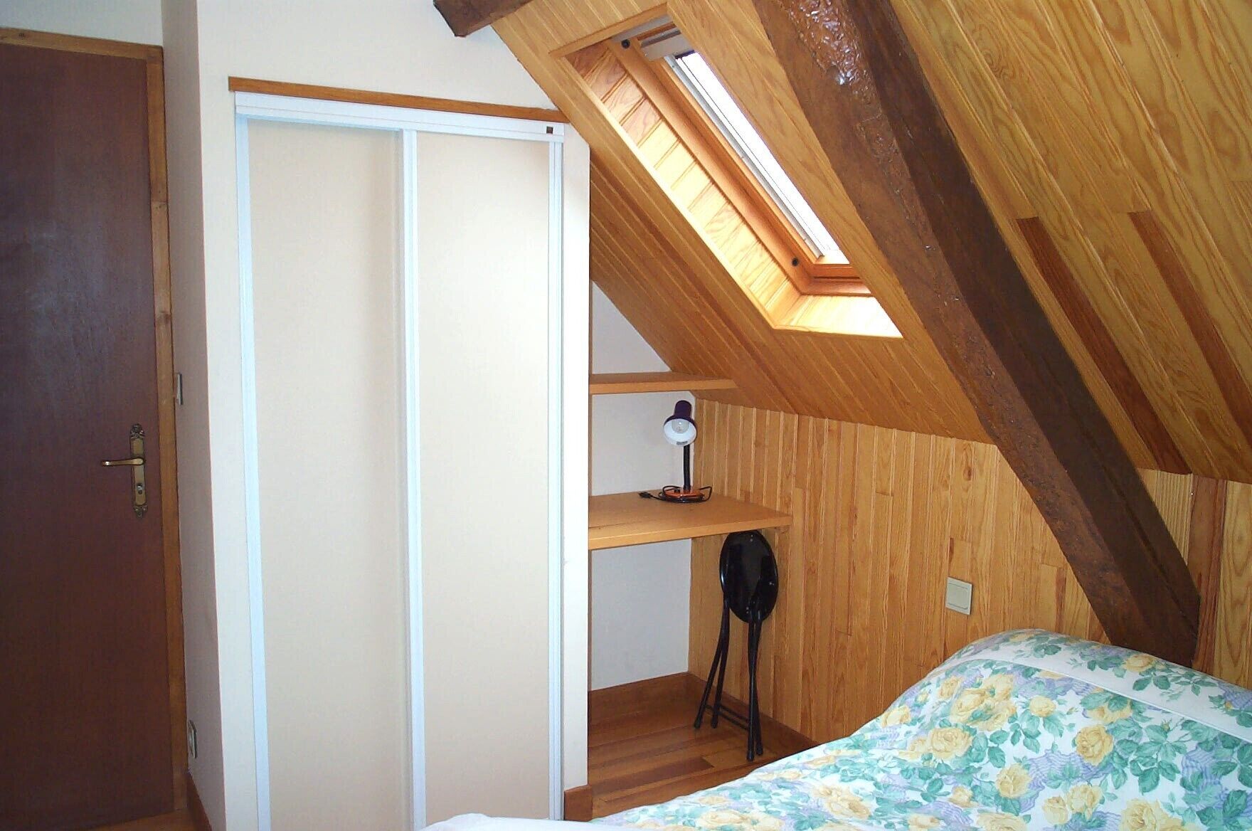 Ein Schlafzimmer mit Oberlicht, einem Bett mit floralem Bettbezug, einem Schreibtisch mit Lampe und einem Kleiderschrank.
