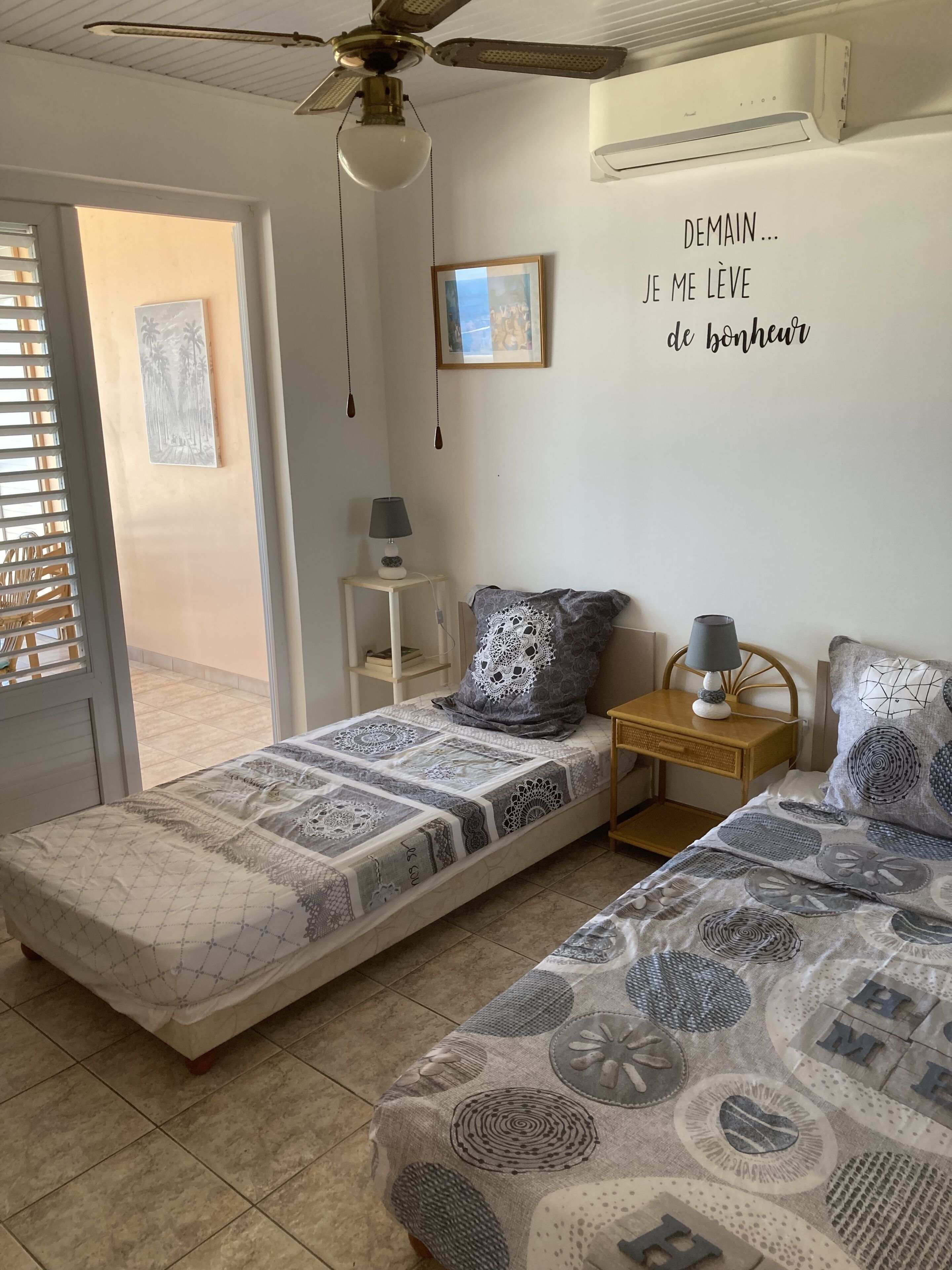 A bedroom with two single beds, a ceiling fan, a small bedside table with a lamp, and a wall decoration with French text.