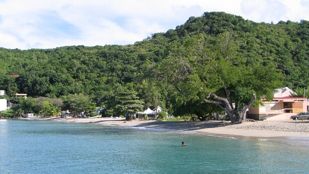A beach with clear blue water, a person swimming, and a hill with trees in the background. - Exceptional panoramic view and proximity to the beach (97217 Les Anses d'Arlet)