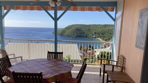 A covered outdoor seating area with a table and chairs, overlooking a coastal view with boats and a green hillside. - Exceptional panoramic view and proximity to the beach (97217 Les Anses d'Arlet)