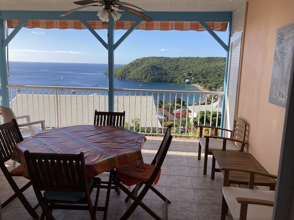 A covered outdoor seating area with a table and chairs, overlooking a coastal view with boats and a green hillside. - Exceptional panoramic views and proximity to the beach (97217 Les Anses d'Arlet)