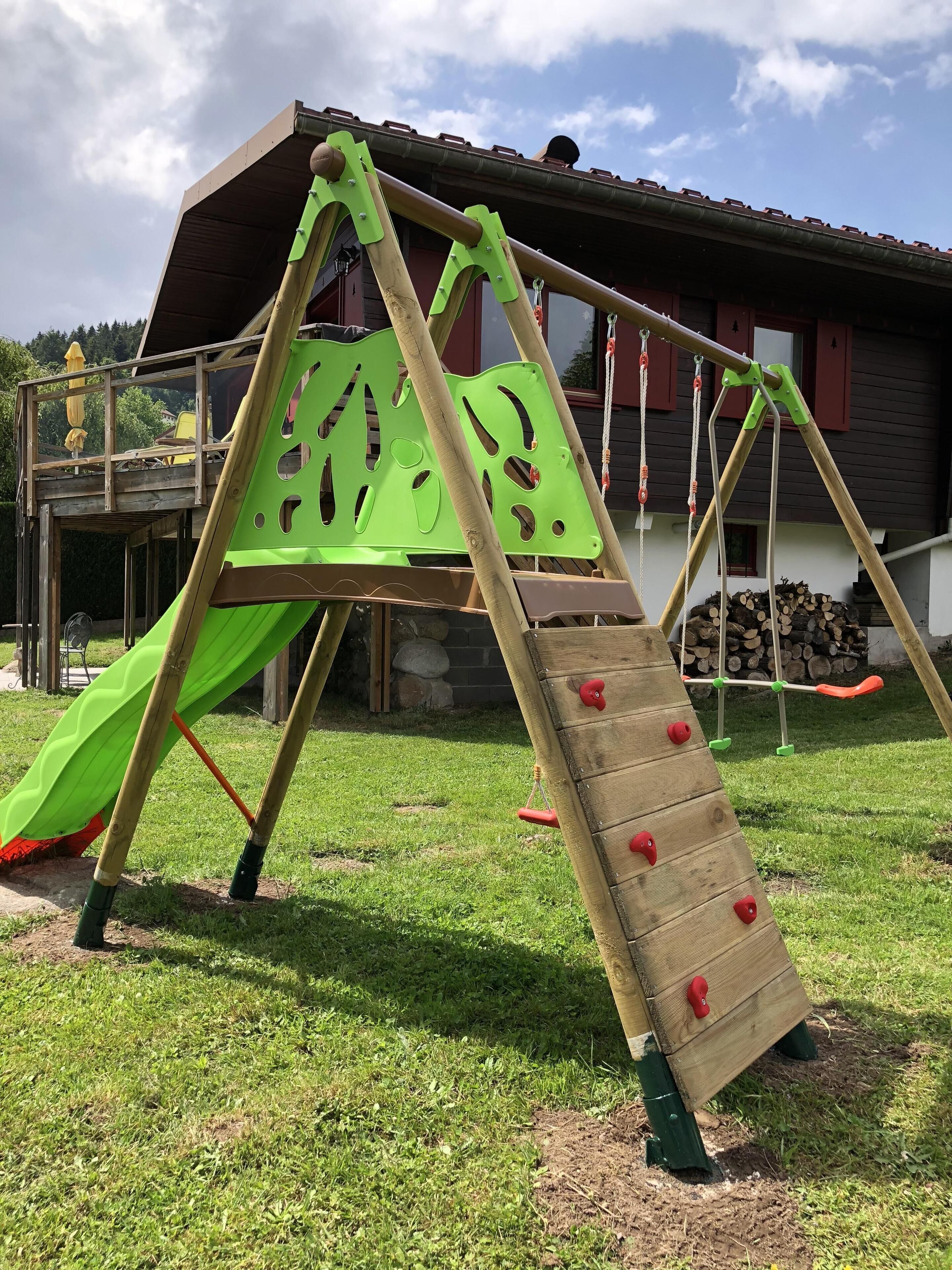Un parque infantil con un tobogán y pared de escalar, frente a una casa con techo rojo y una pila de leña al fondo.