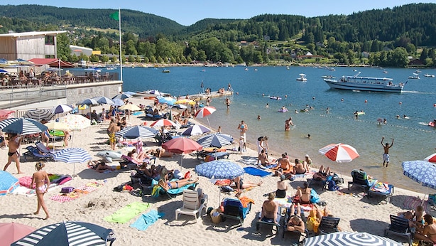 En strand med folk, der solbader og svømmer, parasoller og både på vandet.