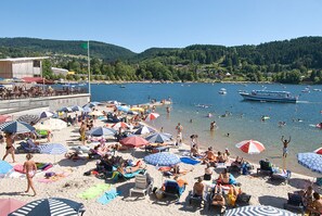 A beach with people sunbathing and swimming, umbrellas, and boats on the water.