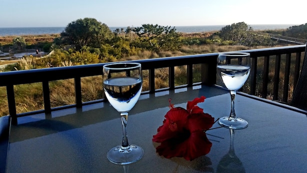 Un tavolo con due bicchieri da vino e un fiore rosso su un balcone affacciato su un paesaggio costiero.