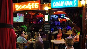 Un área de bar al aire libre con letreros de neón que dicen 'BOOGIE' y 'COCKTAILS'. Hay mesas y sillas para los clientes, y una pantalla de televisión mostrando un evento deportivo.